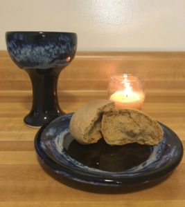 broken whole wheat roll on blue ceramic plate, matching chalice, and burning beeswax vigil candle sitting on counter